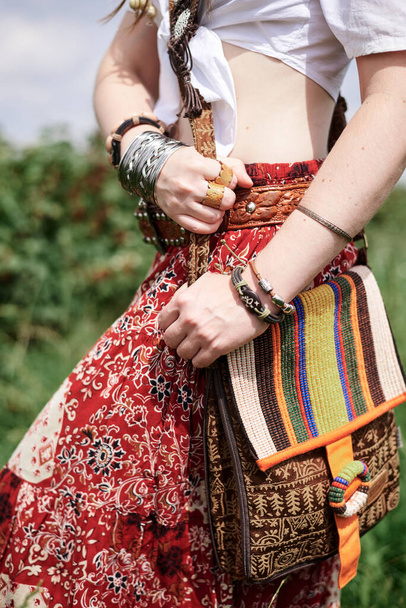 Close-up picture of female hands with bracelets and rings, holding shoulder purse. Young hippie woman, wearing colorful boho style clothes, standing on green field. Eco tourism concept. - Photo, Image