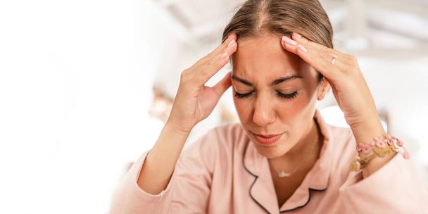 Młoda biała kobieta w różowej piżamie trzyma głowę w dłoniach i grymas w bólu od migreny. - Zdjęcie, obraz