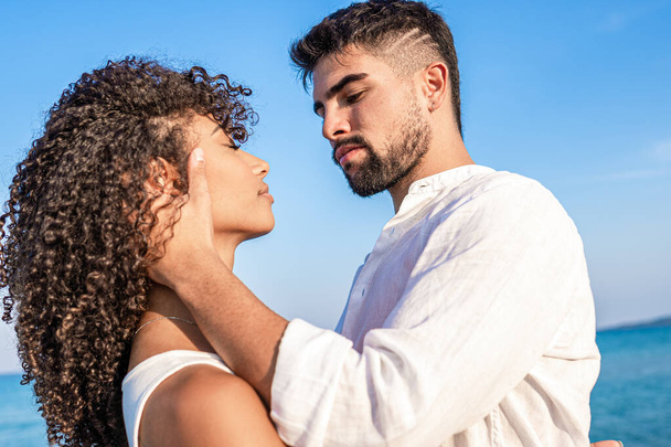 Young handsome bearded man in white shirt grabs his curly hispanic woman forcefully holding her head in his hands with serious expression - Bright and vivid color low angle view - Photo, Image