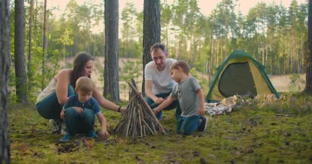 A család egy tábortűz körül üldögél egy nyári estén. A gyerekek a szüleikkel az erdőben pihennek. Hétvége a természetben jó társaságban. A család kolbászt süt egy erdőtűz felett.. - Felvétel, videó