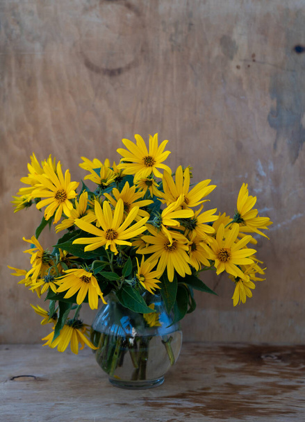 A bouquet of yellow Jerusalem artichoke flowers in a glass vase on a wooden background. Postcard with place for text. A simple wild flower that looks like a chamomile. - Zdjęcie, obraz