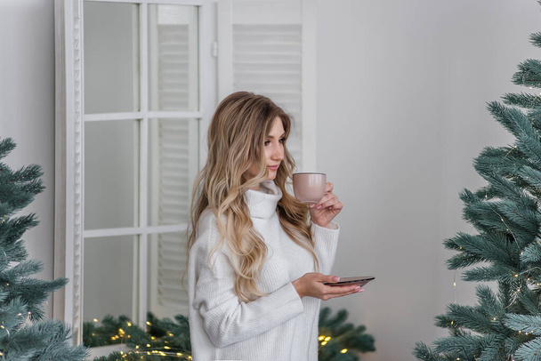 A happy girl in a warm winter sweater stands in a Scandinavian-style kitchen and drinks a hot drink from a pink cup by the Christmas tree. New Year's morning. Festive interior and home decoration. - Fotografie, Obrázek