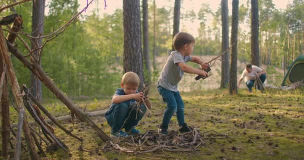 Dva chlapci sbírají v lese klacky pro velký rodinný táborák na pozadí stanu a jezera. Rodina zapálí oheň pro kempování a smažení marshmallows - Záběry, video