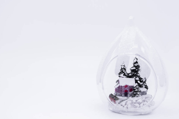 Transparente Weihnachtskugel, in deren Inneren die Figur eines Hauses im verschneiten Wald zwischen den Tannen steht. Weißer Hintergrund, Kopierraum. Gemütliches Winterferienkonzept. - Foto, Bild
