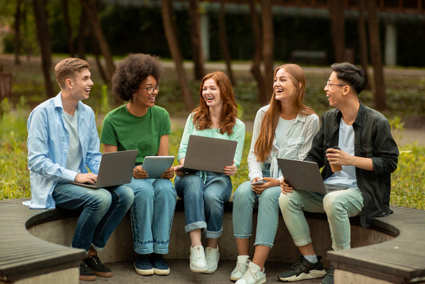 Διάφοροι πολυπολιτισμικοί φοιτητές που μελετούν σε εξωτερικούς χώρους με διαφορετικές ηλεκτρονικές συσκευές, καθισμένοι στο πάγκο - Φωτογραφία, εικόνα