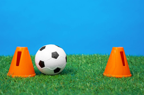 Two plastic cone restraints stand depicting a soccer goal, a small toy football ball between them on the green grass of artificial lawn, blue background, front view, close up - Photo, Image