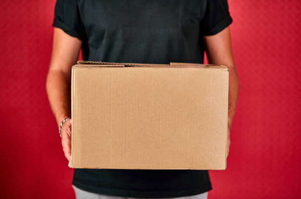 Delivery man employee in black uniform hold empty cardboard box isolated on red background studio, Service quarantine pandemic coronavirus, Contactless delivery. - Valokuva, kuva
