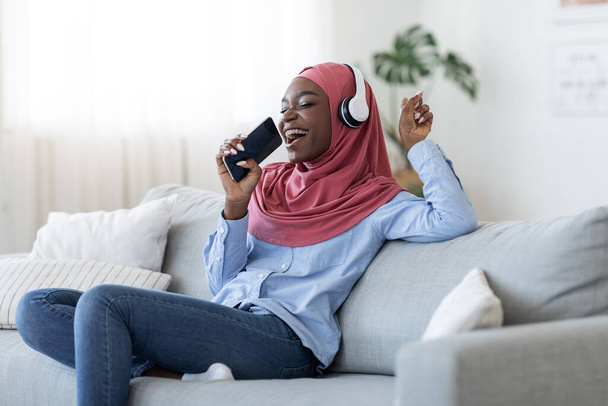 Мобільний караоке. Радісна чорна мусульманка співає зі смартфоном вдома - Фото, зображення