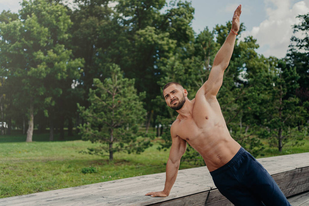Strong young man stands in side plank on one arm, finds balance, poses at park near trees, practices yoga outdoor, leads active healthy lifestyle, has muscualr strong body. Motivated bodybuilder - Foto, afbeelding