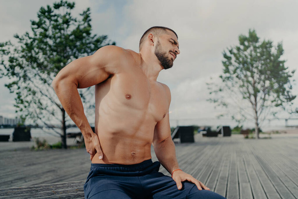 Displeased unshaven European sportsman touches lower back and feels backache after physical training exercises, poses outdoor. Sport exercising injury. Athletic guy suffers pain, needs massage - Fotoğraf, Görsel