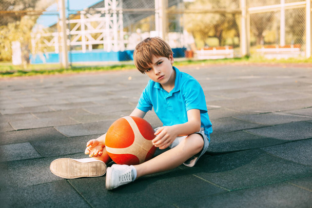 Красивий хлопчик баскетболіст у синій футболці сидить на баскетбольному майданчику з м'ячем у руках. Спортсмен відпочиває після тренування
. - Фото, зображення