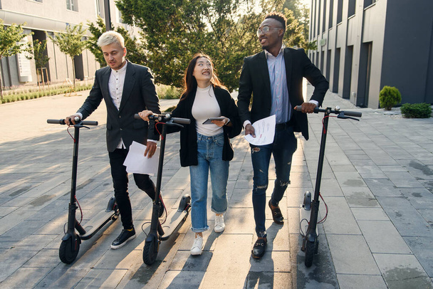 Confident happy multiracial office colleagues discussing business project go near office building with electric scooters - Fotografie, Obrázek
