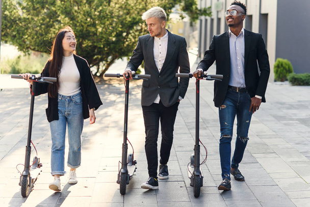 Confident happy multiracial office colleagues discussing business project go near office building with electric scooters - Foto, Imagem