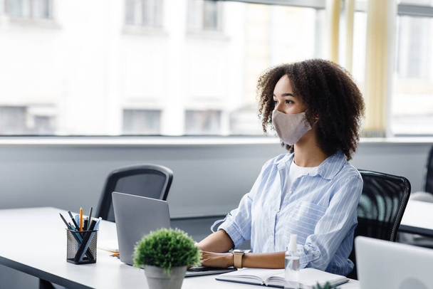 Współpracujące miejsce do pracy. Afroamerykanka w masce ochronnej patrzy na klienta lub kolegę i pracuje w laptopie, siedzi przy stole z laptopem - Zdjęcie, obraz