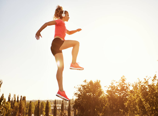 De baixo de corpo inteiro de fêmea esportiva jovem ativa em activewear e fones de ouvido pulando alto contra céu azul sem nuvens durante o treino ao ar livre no verão da - Foto, Imagem