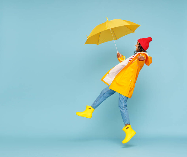 外衣姿の幸せな民族女性の側面図と傘の跳躍と青の背景に雨の秋の日に楽しみを持っている - 写真・画像