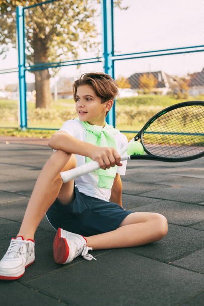 Kauniin teinipojan muotokuva tennismaila kädessään. Tennispelaaja istuu kentällä ja lepää harjoittelun jälkeen. Urheilu, urheilija - Valokuva, kuva