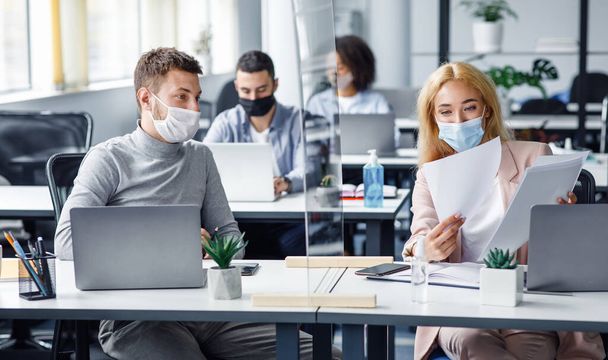 Social distancing and document management. Millennial guy and woman in protective masks discuss work issues through protective board - Photo, Image