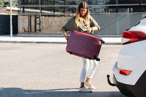 Full body positive young woman smiling and putting luggage into trunk of white vehicle on city street during summer vacation - Photo, Image