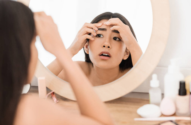 Järkyttynyt aasialainen nuori nainen tarkistaa näppylöitä, katsot peiliin - Valokuva, kuva