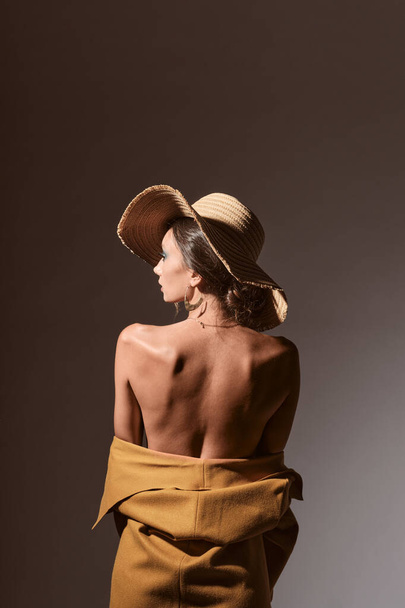 Sexy brunette young woman portrait showing her back and shoulders wearing a yellow coat with a hat - Foto, imagen