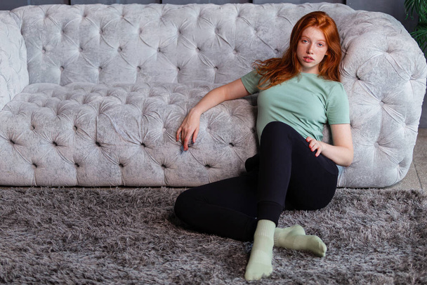 Portret pięknej rudej dziewczyny siedzącej w luźnych ciuchach na podłodze przy kanapie spędzającej czas w salonie w domu. Odpoczynek relaks dobry nastrój rekreacja styl życia koncepcja. - Zdjęcie, obraz