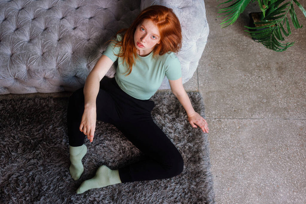 Portrait of a beautiful redhead girl sitting in casual clothes on floor near couch spending time in living room at home. Rest relax good mood leisure lifestyle concept. - Foto, imagen