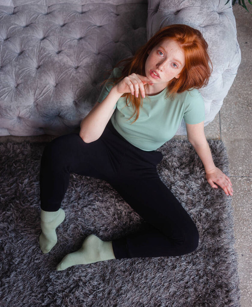 Portrait of a beautiful redhead girl sitting in casual clothes on floor near couch spending time in living room at home. Rest relax good mood leisure lifestyle concept. - Foto, Imagen