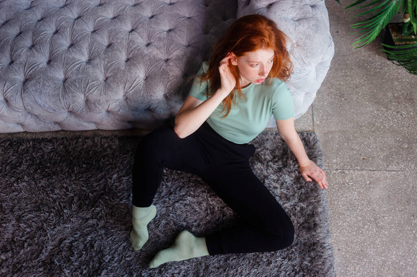 Portrait of a beautiful redhead girl sitting in casual clothes on floor near couch spending time in living room at home. Rest relax good mood leisure lifestyle concept. - Foto, Imagem