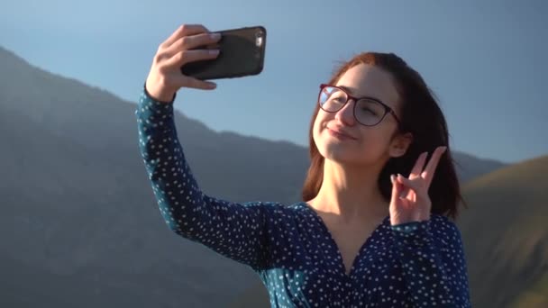 Μια νεαρή γυναίκα με φόρεμα στέκεται στα βουνά και βγάζει μια σέλφι σε ένα smartphone.. - Πλάνα, βίντεο
