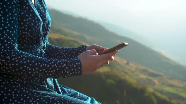 Een jonge vrouw in een jurk zit met een telefoon in haar handen tegen een achtergrond van bergen close-up. Het meisje reist door de Kaukasus. - Video