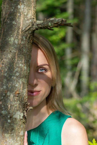 Blonde woman with green blouse hiding behind the tree trunk in the forest. Tree casting the shadow on her face - Fotografie, Obrázek