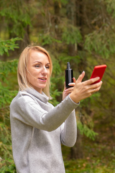 Blonde woman with grey blouse make live stream advertisement on social media about spray in black bottle with red cell phone in dark forest - Фото, зображення