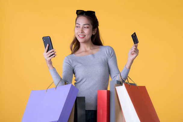 Beautiful young Asian woman with colourful shopping bags with smart phone and credit card isolated on yellow background - 写真・画像