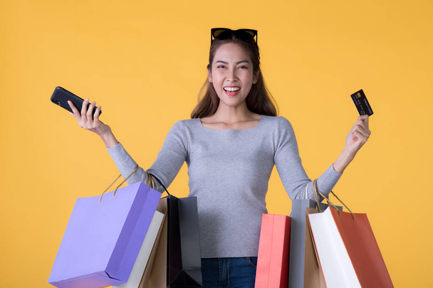Beautiful young Asian woman with colourful shopping bags with smart phone and credit card isolated on yellow background - Фото, изображение