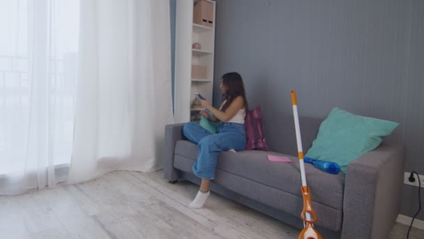 Cansada jovem mulher depois de limpar a casa sozinha senta-se no treinador e usa telefone - Filmagem, Vídeo