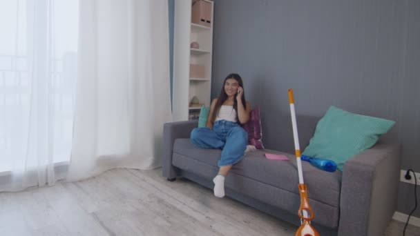 Cansada jovem mulher depois de limpar a casa sozinha senta-se no treinador e usa telefone - Filmagem, Vídeo