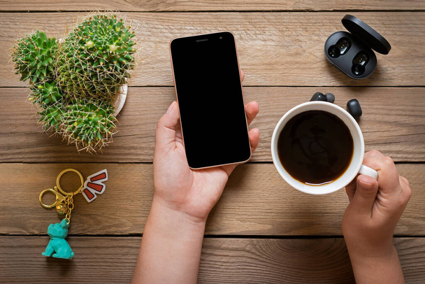 Okostelefon fekete képernyős kijelzővel női kezekben, vezeték nélküli fejhallgató, kaktusz, töltőkapszula, csésze kávé fa asztalon Lapos fektetés Top view Social media, digitális marketing koncepció. - Fotó, kép
