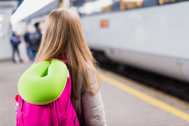 Το κορίτσι περιμένει το τρένο. Κορίτσι με σακίδιο πλάτης και πράσινο μαξιλάρι λαιμού - Φωτογραφία, εικόνα