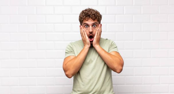 young man feeling shocked and scared, looking terrified with open mouth and hands on cheeks - 写真・画像