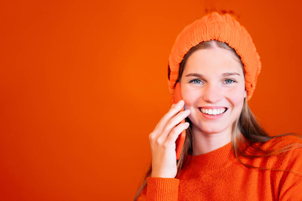 uśmiechnięta piękna kobieta wykonująca telefon - szczęśliwa dziewczyna nosząca wełnianą czapkę na pomarańczowym tle. - Zdjęcie, obraz