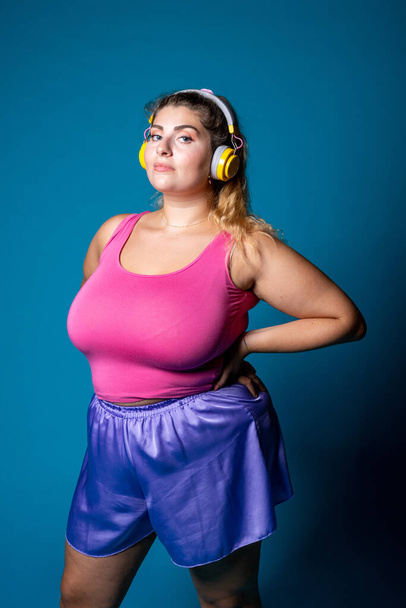 Plus size young black woman posing on blue background listening music - Isolated diverse caucasian female sportive wearing headphones - body positive, training, sport concept - Fotó, kép