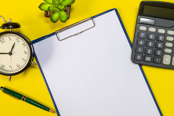 Stationery or essential office tools. Workplace pen with sheets of office paper, calculator, flower and alarm clock close-up on yellow background, top view. - Photo, Image