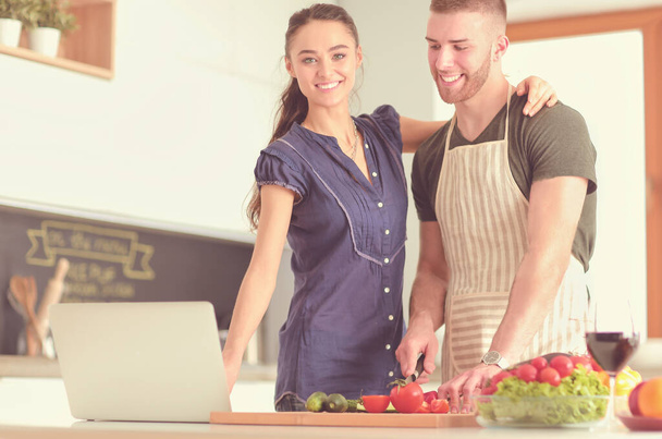 Young man cutting vegetables and woman standing with laptop in the kitchen - Foto, Imagem