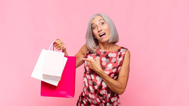 жінка середнього віку виглядає схвильованою і здивованою, вказуючи на бік і вгору, щоб копіювати простір з сумками
 - Фото, зображення