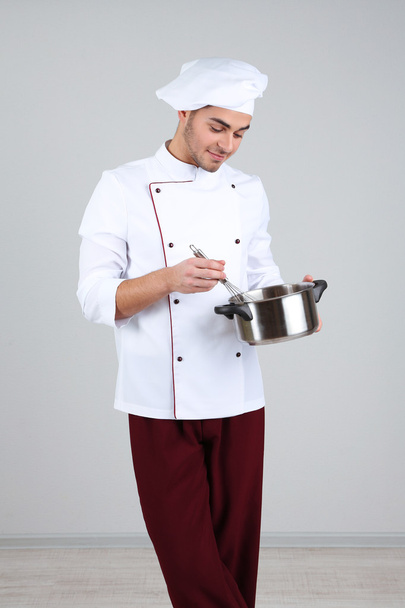 Professional chef in white uniform and hat, holding pan in his hands, on gray background - Foto, afbeelding