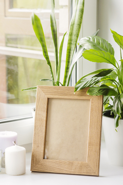A wooden frame stands on the windowsill next to candles and home flowers. Mockup for a wooden photo frame. Poster mockup. Clean, modern, minimal frame. Empty frame Indoor interior,show text or product - Φωτογραφία, εικόνα