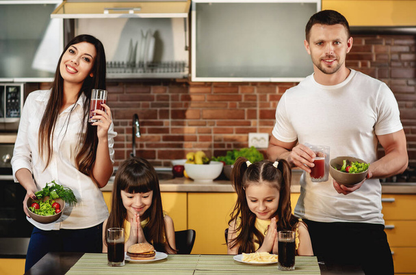 Alimentación saludable frente a malos hábitos. Los padres ofrecen a sus hijas una ensalada con jugo en lugar de una hamburguesa y refresco. Las niñas no son felices. Concepto de comida chatarra - Foto, imagen