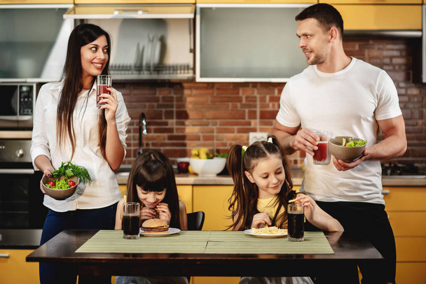Υγιεινή διατροφή εναντίον κακές συνήθειες. Οι γονείς προσφέρουν στις κόρες τους μια σαλάτα με χυμό αντί για μπέργκερ και σόδα. Τα κοριτσάκια δεν είναι χαρούμενα. Έννοια πρόχειρου φαγητού - Φωτογραφία, εικόνα