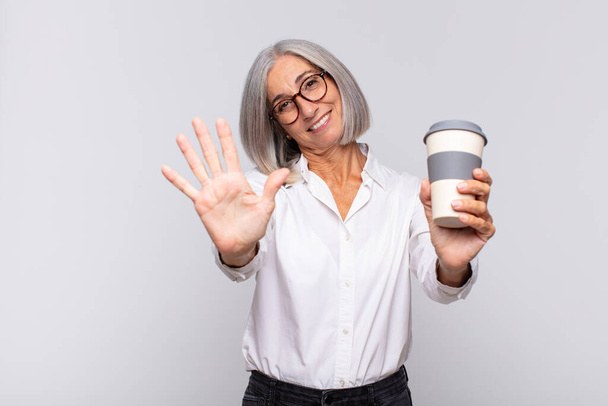žena středního věku se usmívá a vypadá přátelsky, ukazuje číslo pět nebo pět s rukou dopředu, odpočítávání konceptu kávy - Fotografie, Obrázek
